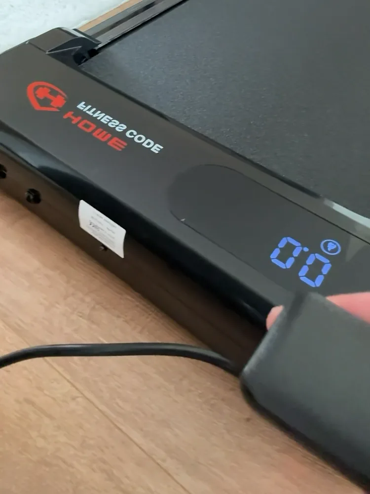 Home Fitness Code walking pad unpacked on a wooden floor, plugged in with its blue display lit